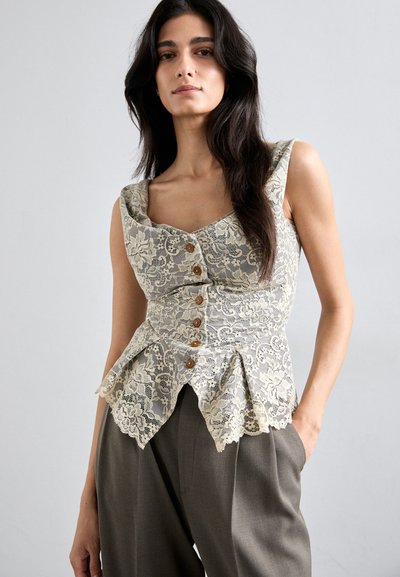 Kerman peplum-topp i krem og grått med hjerteformet utringning, fem brune knapper og bølget kant. Båret sammen med løse grå bukser.