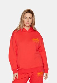 Felpa rossa con cappuccio e tasca frontale, con testo grafico giallo sul petto, abbinata a pantaloni della tuta, realizzata in tessuto morbido.