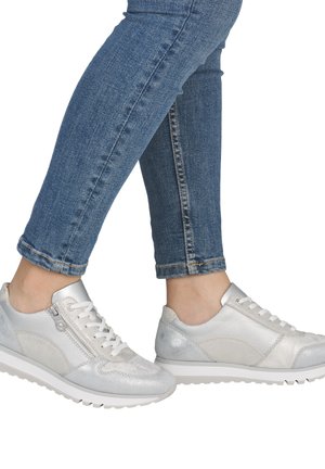 Jambes portant un jean skinny bleu et des baskets argentées à tige basse avec des lacets et des fermetures éclair latérales, vues de côté et de face.