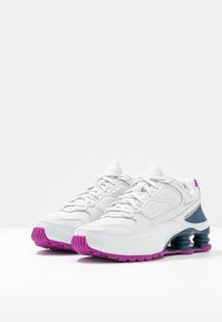 Bílé tenisky s texturovaným koženým a síťovaným svrškem, doplněné fialovou a černou podrážkou, s polstrovaným designem a tkaničkami.