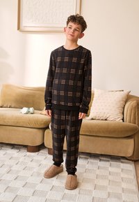 Sort og brun ternet pyjamassæt lavet af blødt stof, lange ærmer, elastisk talje, kombineret med beige slip-on hjemmesko.