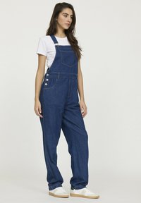 Salopette en denim bleu foncé avec une coupe loose, dotée de bretelles réglables, d'une poche avant et de détails en boutons sur les côtés. Portée sur un T-shirt blanc.