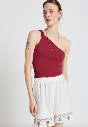 Donna che indossa un top bordeaux monospalla senza maniche e pantaloncini bianchi con ricamo di palme, in piedi davanti a uno sfondo semplice.