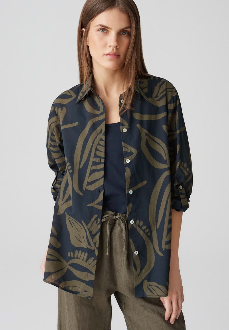 Camicia in tessuto blu navy con motivo a foglie verdi, con chiusura a bottoni, maniche lunghe e vestibilità comoda, abbinata a pantaloni scuri.