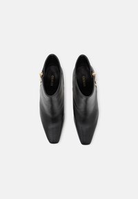 Botines de cuero negro con punta puntiaguda, textura suave y detalle de cremalleras doradas en el lateral. Nombre de la marca "Guess" visible en el interior.