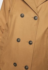 Hnědý dvousečný kabát s černými knoflíky, viditelnou boční kapsou a podrobným prošíváním na hladkém materiálu.