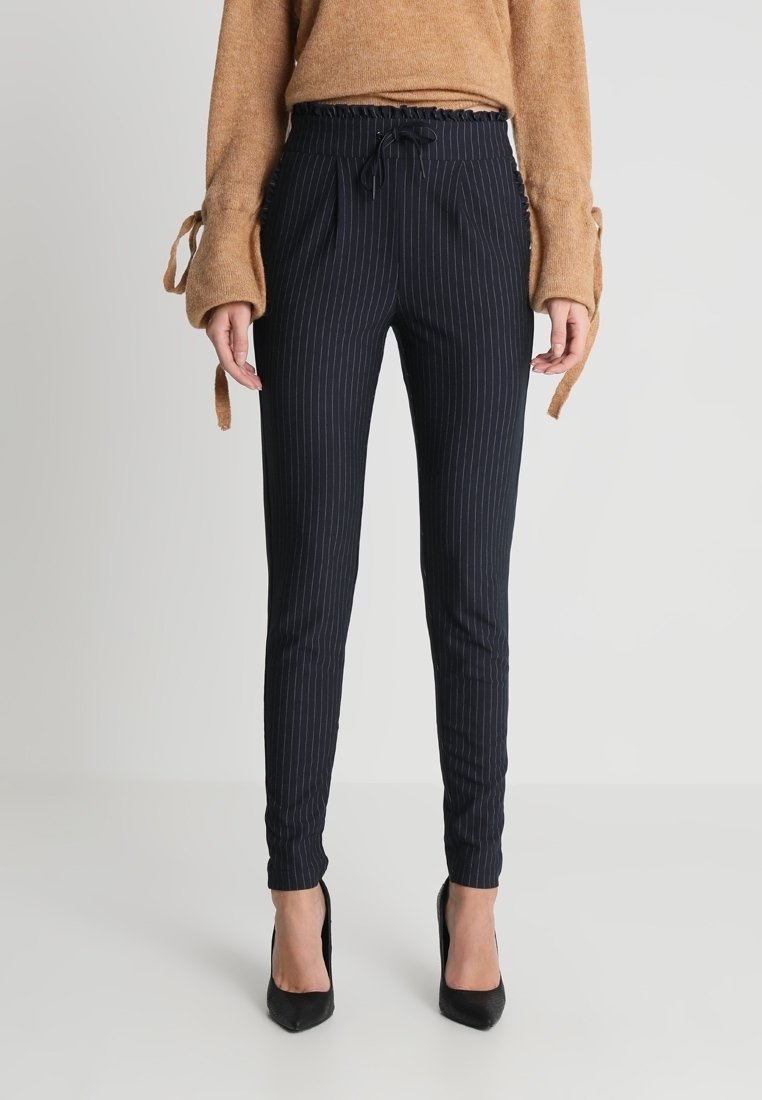 Femme portant un pantalon slim noir à fines rayures avec une taille à cordon, un pull marron à manches longues et des talons hauts noirs, debout droite.