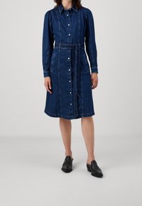 Jeansskjol med lång ärm, knäppning framtill och midjebälte. Har sömdetaljer och fickor. Mörkblå färg.