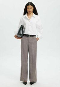 Weißes Langarmshirt mit Knöpfen, graue Karohosen, schwarzer Gürtel und spitze schwarze Absätze. Hält eine schwarze Tasche mit strukturierter Oberfläche.