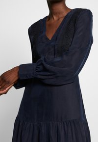 Marinblå klänning med långa ärmar, med V-ringning, spetsdetaljer på axlarna och en veckad kjol. Slät tyg med en lätt lyster.