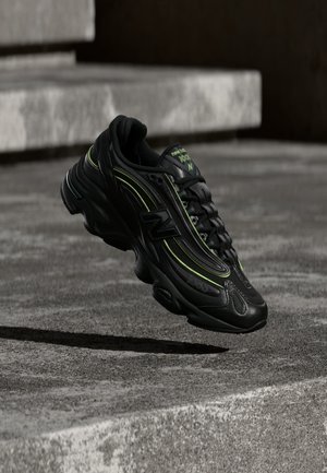 Chaussure de sport noire New Balance avec des accents vert fluo suspendue au-dessus de marches en béton gris.