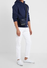 Borsa a tracolla blu navy con due scomparti con zip e logo "POLO SPORT". Indossata da un modello con jeans bianchi e scarpe da ginnastica bianche.