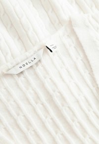 Țesătură albă tricotată cu textură verticală ridicată, tip coaste. Eticheta prezintă "NOELLA" și mărimea S, cu o etichetă suplimentară care indică "Produs în Italia".