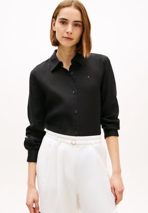 Kvinde iført en sort, langærmet skjorte med knapper, proppet ned i hvide bukser med høj talje og et tyndt hvidt bælte.