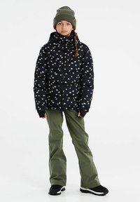 Zwarte pufferjack met een wit bloemenpatroon, hoge kraag, gecombineerd met olijfgroene broek en een gebreide olijfgroene beanie, staande op zwarte schoeisel.