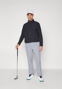 Γκρι παντελόνι γκολφ σε συνδυασμό με μαύρο πουλόβερ με φερμουάρ στο ένα τέταρτο και λευκά παπούτσια. Μια λευκή καπέλο ολοκληρώνει το ντύσιμο. Ο παίκτης γκολφ κρατάει ένα putter.