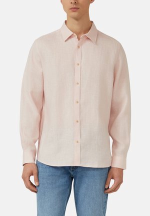 Mann trägt ein hellrosa langärmeliges Leinenhemd mit beigen Knöpfen, kombiniert mit blauen Jeans, vor einfarbigem Hintergrund.