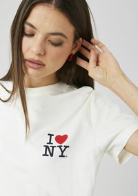 T-shirt en coton blanc arborant un texte noir « NY » avec une icône de cœur rouge. Manches courtes et encolure ronde classique. Design simple.