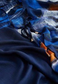 Eșarfă albastră vaporoasă, cu un amestec de modele abstracte în nuanțe de bleumarin închis și albastru deschis, cu accente legate și detalii aurii din metal.