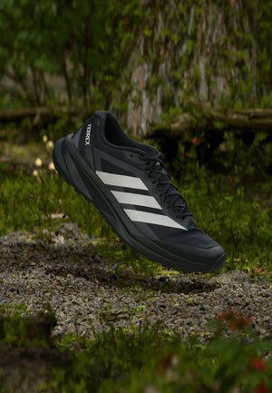 Sort Adidas Terrex trail løbesko med hvide striber, ophængt over en klippefyldt skovbund med grøn bevoksning i baggrunden.