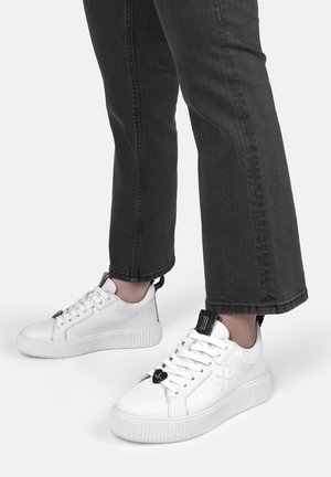 Weiße Leder-Sneaker mit strukturierten Oberflächen, schwarzen Akzenten an der Zunge und einem herzförmigen Schnürdetail. Flache Gummisohle und runde Form.