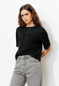Pull noir à manches courtes avec une texture douce et un léger éclat, associé à un jean gris orné de clous métalliques.