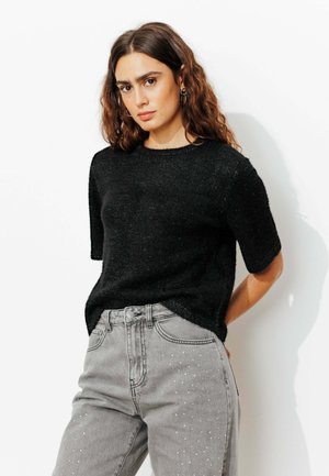 Pull noir à manches courtes avec une texture douce et un léger éclat, associé à un jean gris orné de clous métalliques.