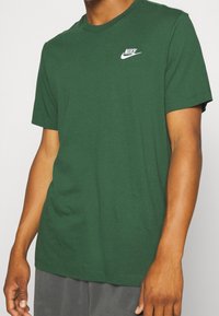 Muž v tmavě zeleném tričku Nike bez potisku s bílým logem na hrudi, v šedých kalhotách, na jednobarevném pozadí.