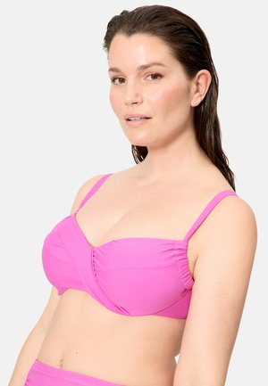 Mujer con el cabello mojado, vestida con un top de bikini rosa brillante, posando contra un fondo liso de color gris claro, mirando ligeramente hacia la derecha.