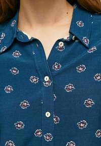 Blå skjorta med knappar i tyg med blommönster i röd och vit, med en vågig krage och krämfärgade knappar.