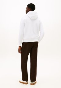 Valkoinen huppari, jossa on resorit hihansuissa ja rento istuvuus, yhdistetty ruskeisiin housuihin ja valkoisiin tennareihin, joissa on kumipohja. Minimalistinen muotoilu.