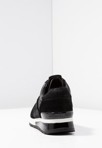 Black Michael Kors sneaker shown from rear with mixed materials and white midsole on white surface and background.