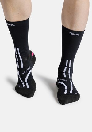 Voeten met zwarte sportsokken met witte en roze grip-patronen, gezien van achter tegen een effen witte achtergrond.