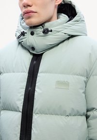 Svijetlozelena puffer jakna s visokim ovratnikom, zatvaranjem na patentni zatvarač, crnim akcentima i logom na prsima. Glatka, podstavljena tekstura.