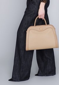 Donna che tiene in mano una borsa a tracolla beige con manici superiori, indossando jeans svasati scuri e scarpe nere a punta su uno sfondo neutro.