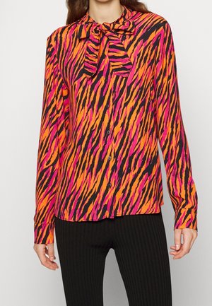 Kobieta ubrana w jaskrawą bluzkę w tygrysie paski w kolorach różowym, pomarańczowym i czarnym, z zapinanym przodem i kokardą przy szyi, w połączeniu z czarnymi spodniami.