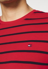 Rød bomuld t-shirt med horisontale sorte striber, rund hals, og et lille broderet logo i nedre højre hjørne.