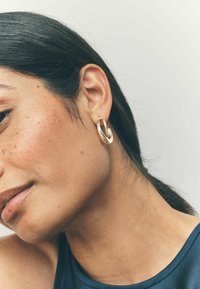 Orecchino a cerchio in oro con una superficie liscia e lucida. Il suo design circolare presenta un accento ondulato, che riflette la luce sul tono della pelle.