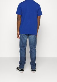 Camiseta de algodón azul, de manga corta, con cuello redondo; combinada con jeans de mezclilla azul, de pierna recta, usados con zapatillas negras con acentos blancos.