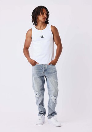 Giovane uomo con dreadlocks che indossa una canotta bianca, jeans chiari strappati e sneakers bianche, in piedi con le mani nelle tasche.