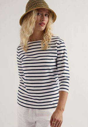 Femeie blondă purtând o pălărie tip găleată tricotată, verde și maro, un tricou cu mânecă lungă cu dungi albastre și albe și pantaloni albi, stând în picioare pe un fundal simplu.