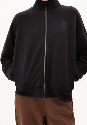 Person trägt eine schwarze Jacke mit Reißverschluss und hohem Kragen sowie kleinem Logo auf der Brust, kombiniert mit brauner Hose, die Hände in den Jackentaschen.