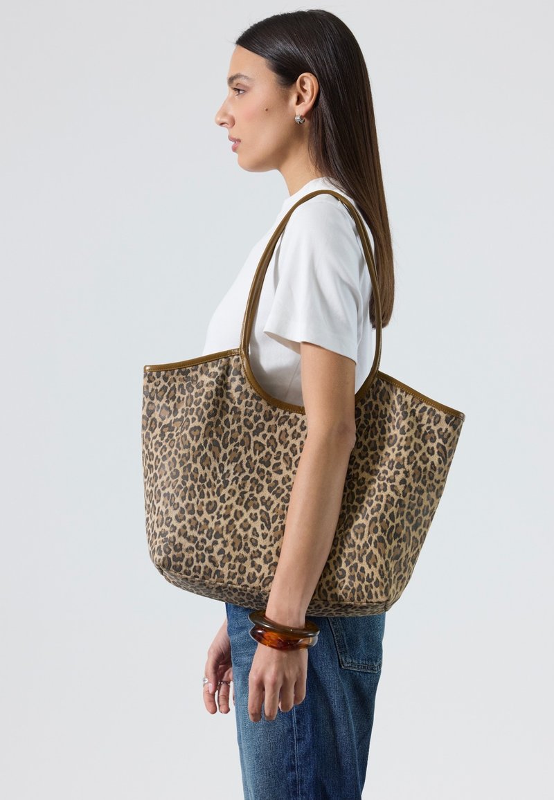 Femme aux cheveux raides et foncés portant une chemise blanche et un jean, portant un grand sac cabas à motif léopard sur l'épaule, sur un fond uni.
