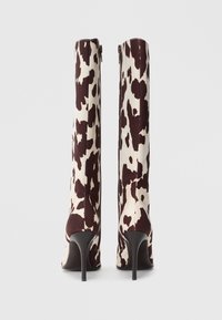 Cizme lungi cu toc, cu un model în crem și imprimeu de vacă maro. Prezintă o siluetă elegantă și închidere cu fermoar pe lateral, având un toc subțire stileto.