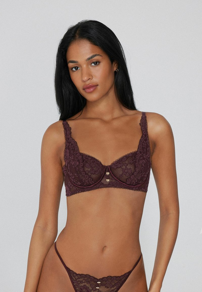 Bordový krajkový podprsenka s květinovým vzorem, s podprsenkovou výztuhou, širokými ramínky a zlatými srdí čkovými akcenty uprostřed. Součástí je tanga.