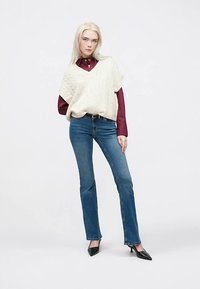 Strikket kremfarget vest med flettemønster over en burgunder langermet skjorte, kombinert med blå vide jeans og svarte spisse hæler.