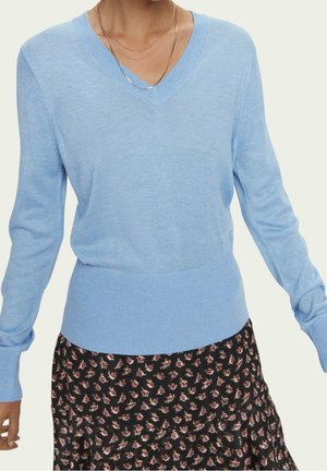 Pull bleu clair en V fabriqué à partir d'un tissu doux, avec un ourlet et des poignets côtelés. Porté par-dessus une jupe noire à motifs floraux.