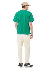 Hombre con camiseta verde, pantalones crema, zapatillas negras y blancas, y una gorra de béisbol beige, de pie de espaldas sobre un fondo blanco.