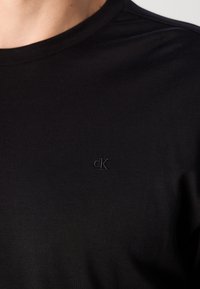 Svart crewneck skjorte med subtilt svart "cK" logo brodert på venstre bryst, båret av en person med lys hudtone.