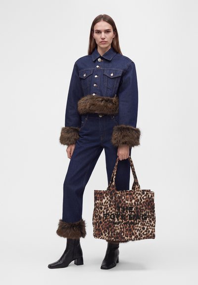 Femme portant une veste en jean bleu foncé et un pantalon avec une bordure en fourrure marron, des bottes noires, tenant un grand sac fourre-tout imprimé léopard avec du texte.